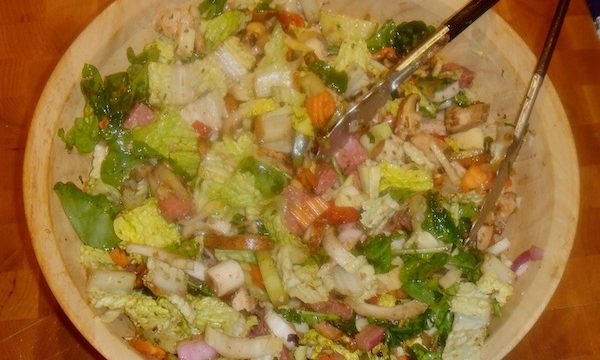 A large light wood salad bowl filled with greens, ham, cheese, tomatoes and carrots.