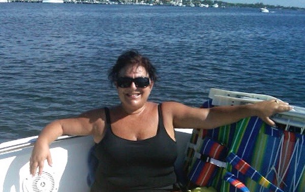 A dark haired woman with sunglasses in a black bathing suit sitting on the back of a boat on the water