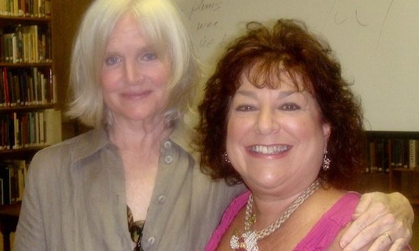 Woman with blond hair and tan suit with her arm around shorter woman with curly dark brown hair in bright pink top