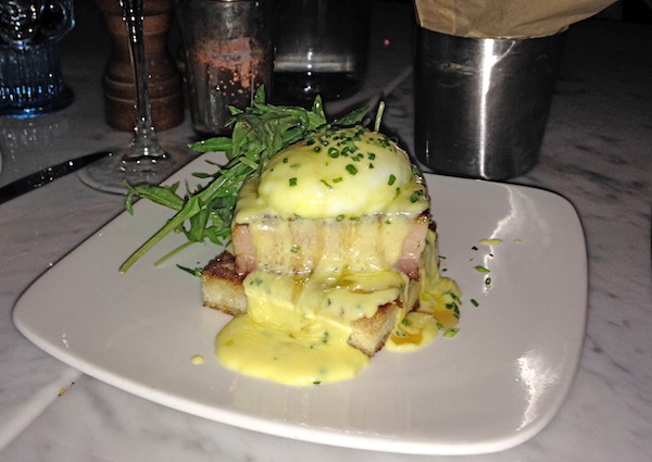 A square white plate topped with pork belly and poached egg with bearnaise sauce dripping down and garnished with chives and greens