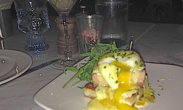 A square white plate topped with pork belly and poached egg with bearnaise sauce and egg yolk dripping down and garnished with chives and greens