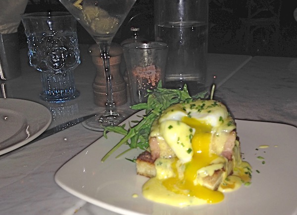 A square white plate topped with pork belly and poached egg with bearnaise sauce and egg yolk dripping down and garnished with chives and greens