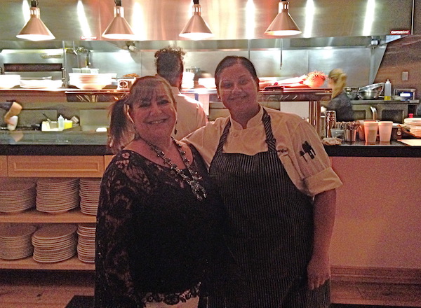A woman in a lace top and a chef with a black apron in front of the kitchen