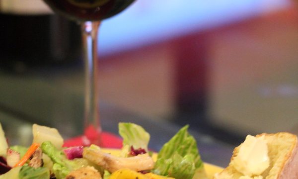 A yellow dinner plate piled high with salad, mango chunks and chicken with a slice of buttered baguette. A glass of red wine is behind the salad