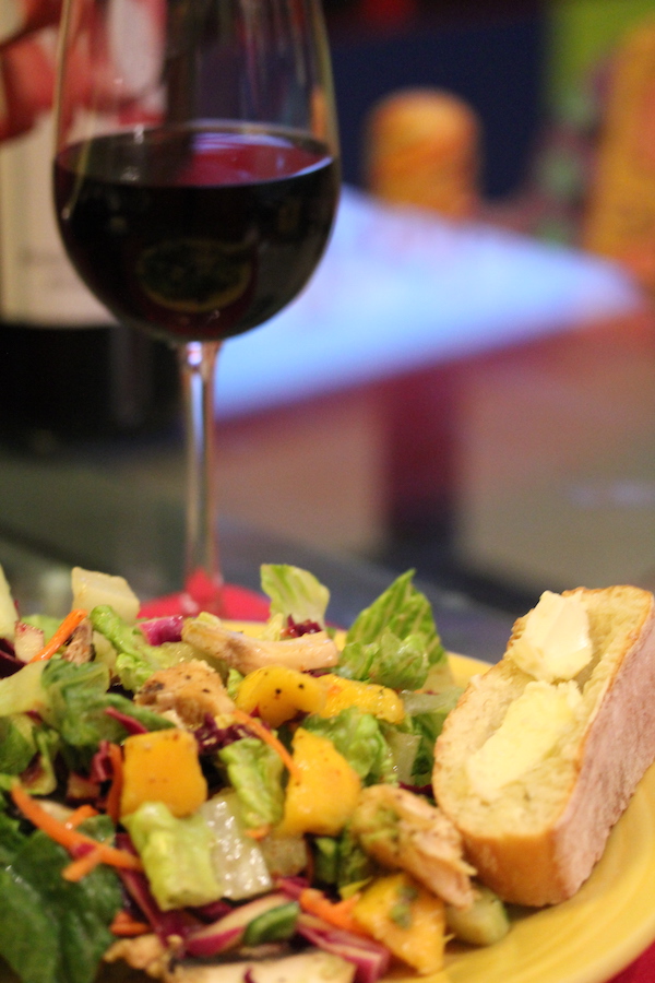 A yellow dinner plate piled high with salad, mango chunks and chicken with a slice of buttered baguette. A glass of red wine is behind the salad