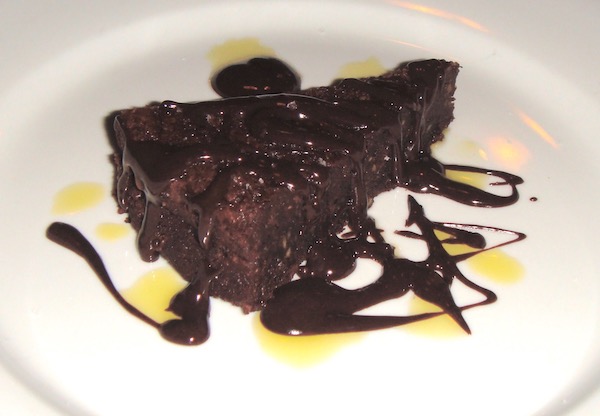 A white plate topped with a wedge of dark chocolate cake drizzled with fudge sauce.
