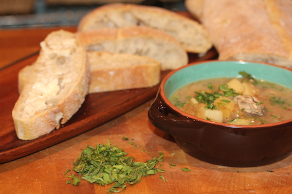 On right a brown crock with turquoise interior filled with an oyster stew sitting on a butcher block. On left is a wooden bread tray with slices of French bread. Between the two items is some chopped parsley directly on the butcher block.