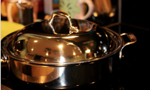 A large stainless steel sauté pan with lid in foreground and colorful ceramics in background.