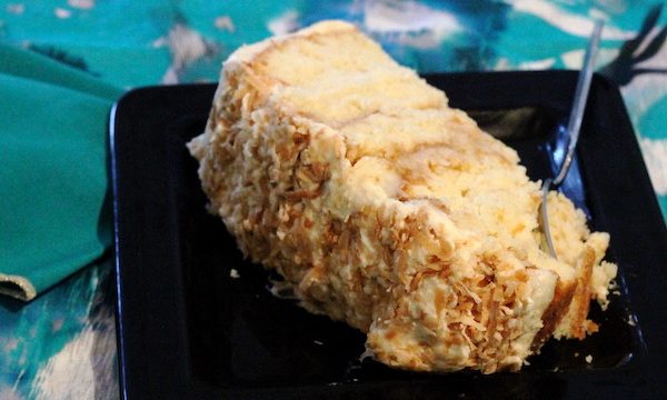 A slice of coconut layer cake on a black plate with a fork sitting on a turquoise print tablecloth.
