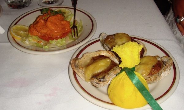 Two plate of food on a white tablecloth. In foreground a trio of oysters with yellow fabric wrapped lemon half. In Background Shrimp with Remoulade Sauce on bed of lettuce with lemon slices