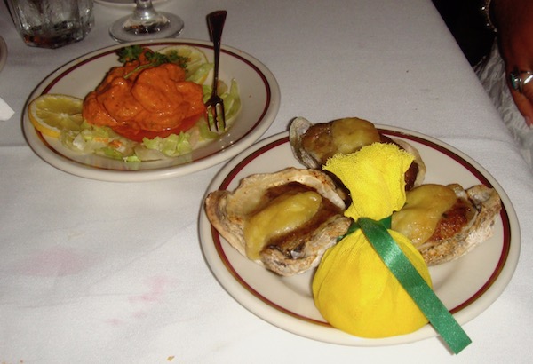 Two plate of food on a white tablecloth. In foreground a trio of oysters with yellow fabric wrapped lemon half. In Background Shrimp with Remoulade Sauce on bed of lettuce with lemon slices