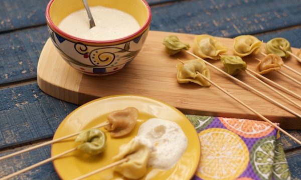 A yellow plate with skewered tortellini and a wood board with more tortellini and a painted ceramic bowl of white sauce with a spoon handle sticking out.