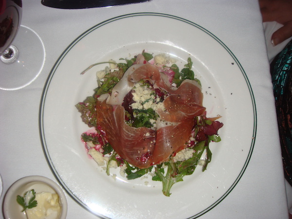 Black rimmed white plate holding and arugula salad topped with thinly sliced prosciutto and crumbled blue cheese