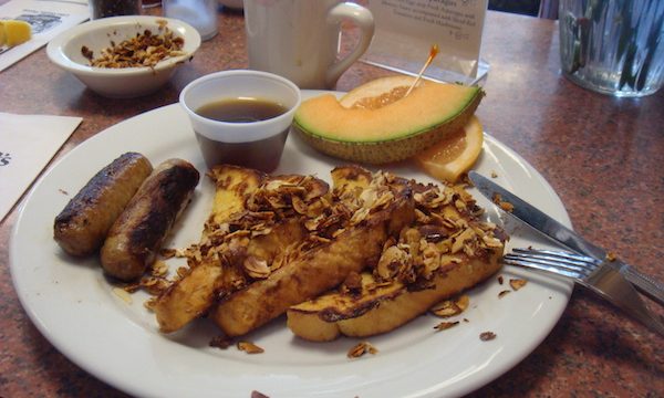 White plate topped with French Toast triangles, almonds, sausages, syrup in a plastic cup and cantaloupe on a wood table