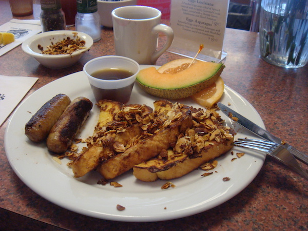 White plate topped with French Toast triangles, almonds, sausages, syrup in a plastic cup and cantaloupe on a wood table