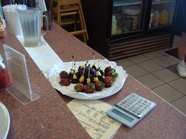 A wood counter with a plate of chocolate dipped fruit, a menu and a calculator.