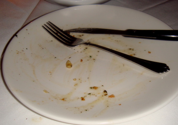 An empty white dinner plate topped with a fork and knife and a few specks of food.