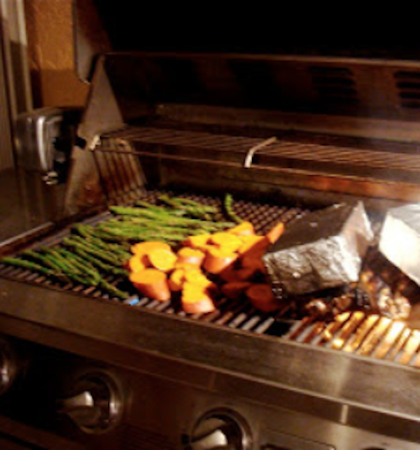 Stainless steel grill with asparagus, foil wrapped bricks atop chicken and additional veggies.