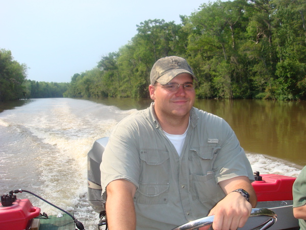 Man in khaki with khaki cap driving a boat in the bayou