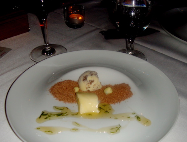 A large white plate on a table set with wine glasses the plate is filled with a scoop of ice cream, a pecan tuile, and a light green sauce flecked with bright green