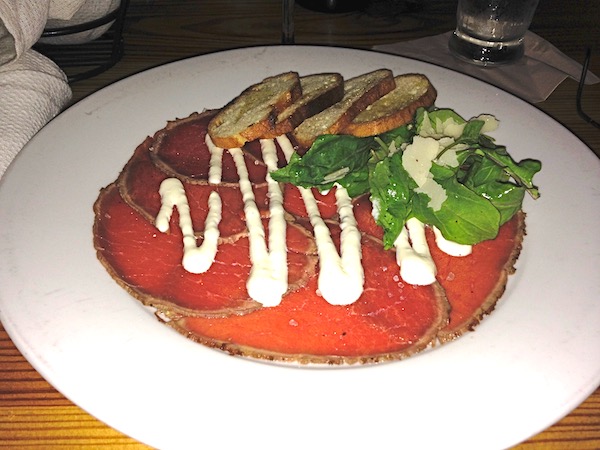 A white plate topped with rare thin slices of beef, toasted baguette slices and a large zigzag of white sauce.