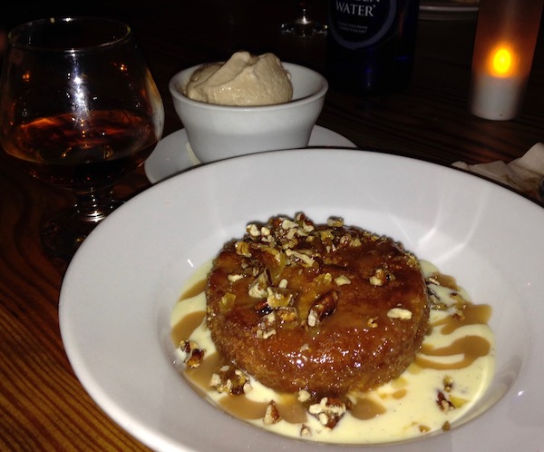 A white shallow bowl in front filled with a small round cake and creamy sauce. In back of that is a small bowl of ice cream