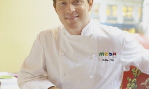 Man with red hair in chef whites sitting at a table with right arm resting on table.