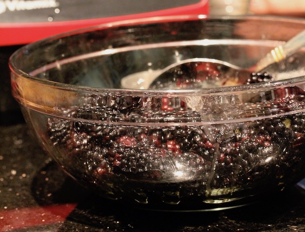 A large glass bowl filled with blackberries being stirred with a stainless serving spoon