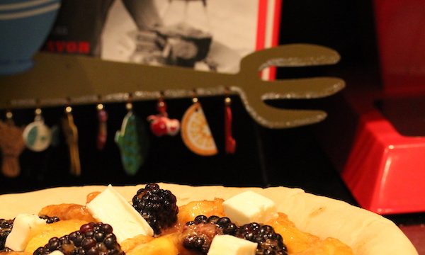 A pie pan with unbaked pie crust draped over it and filled with peaches, blackberries and cubes of butter. A cookbook on a stand is behind pie.