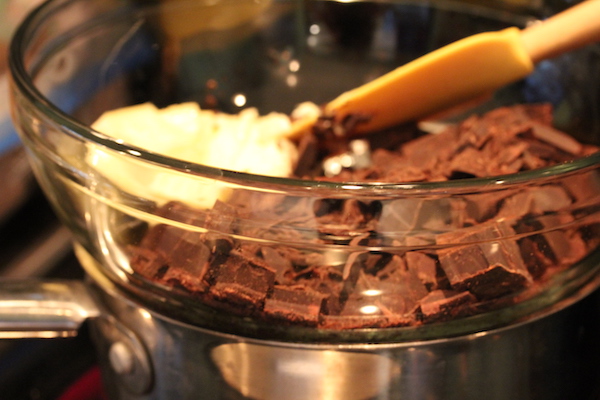 Chocolate chunks and butter in a glass bowl over a saucepan being stirred by a yellow spatula.