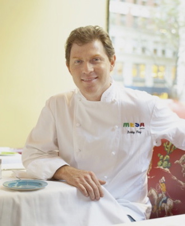 Man with red hair in chef whites sitting at a table with right arm resting on table.