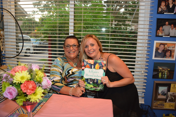 A woman with glasses seated and a woman in a black dress holding a book.
