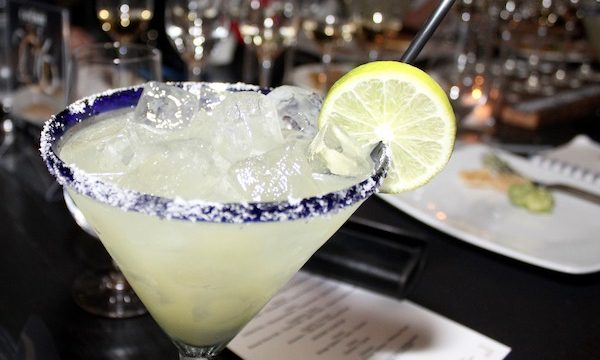 A blue rimmed martini glass with salt on the rim filled with pale yellow liquid and a thin lime wedge on the side of the glass rim.