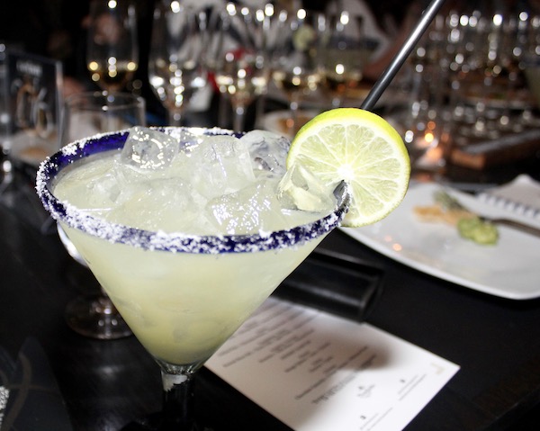 A blue rimmed martini glass with salt on the rim filled with pale yellow liquid and a thin lime wedge on the side of the glass rim.