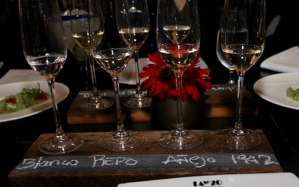 A wood table in a restaurant set with plates and many glasses some filled with tequila 