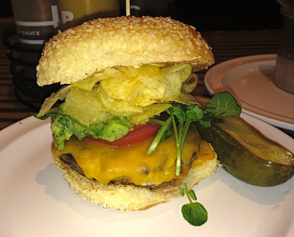 A white plate topped with a huge cheeseburger with watercress and potato chips in a sesame seed bun.
