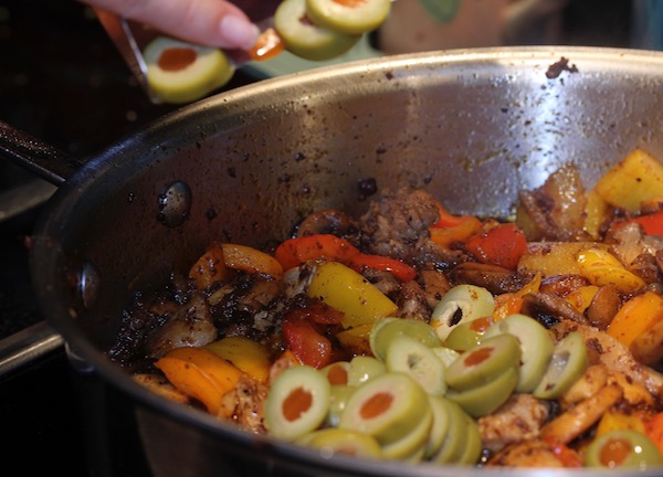 Part of a woman's hand shown adding sliced pimento stuffed olives to a pan of peppers, chorizo and chicken chunks