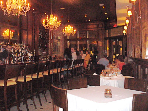 A bar area with dark wood, a long line of bar stools and small tables with white tablecloths.