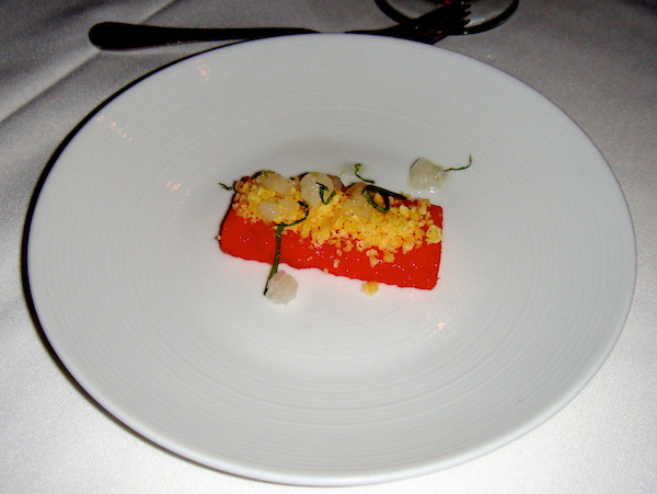 A white plate with a small rectangle of watermelon topped with yellow beads and chiffonade of mint.