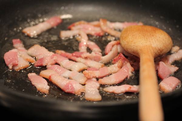 An anodized sauté pan with lardons of bacon cooking and a wooden spoon.