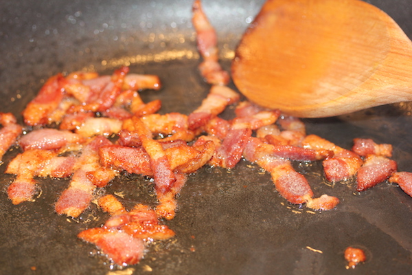 An anodized sauté pan with lardons of bacon cooking to a golden brown and a wooden spoon.