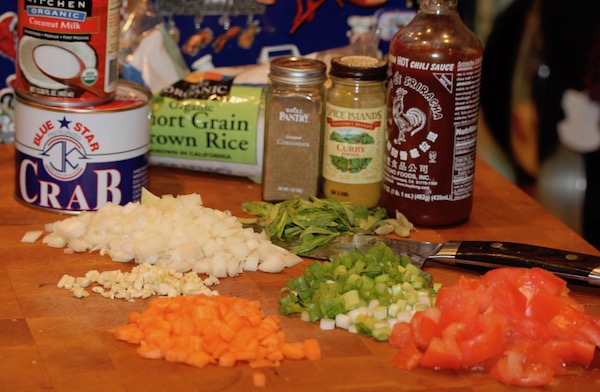 Butcher block with cut carrots, tomatoes, scallions, basil, onions and garlic on it along with a chef's knife, bottle of sriracha, curry powder, ground coriander, cans of crabmeat and coconut milk. 