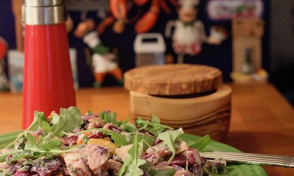 An olive colored dinner plate topped with sausage chunks and greens sitting on a butcher block along with an olive wood salt cellar and a red pepper mill.