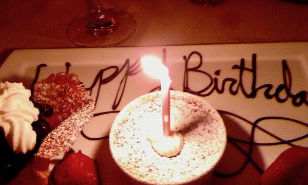 A white rectangular plate with a ramekin filled with custard and a candle, whipped cream, strawberries and Happy Birthday written in chocolate.