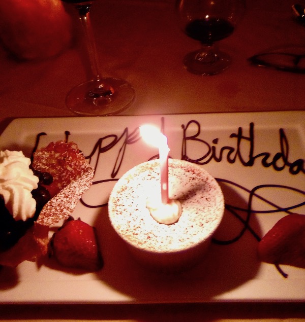 A white rectangular plate with a ramekin filled with custard and a candle, whipped cream, strawberries and Happy Birthday written in chocolate.