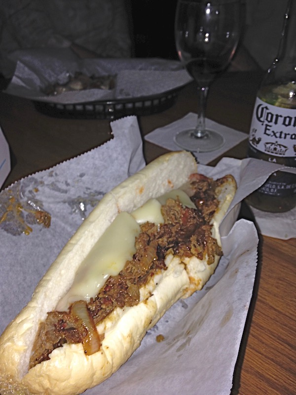 A cheesesteak on white deli paper with a Corona and a glass of red wine in the background