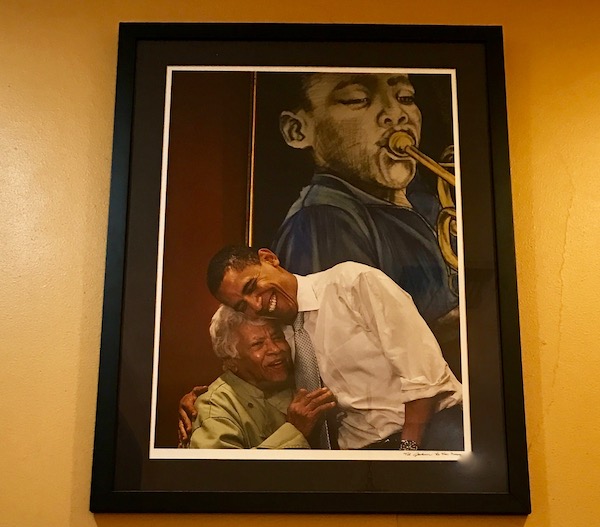 A framed photo of President Obama hugging Chef Leah Chase in front of a painting of a jazz musician