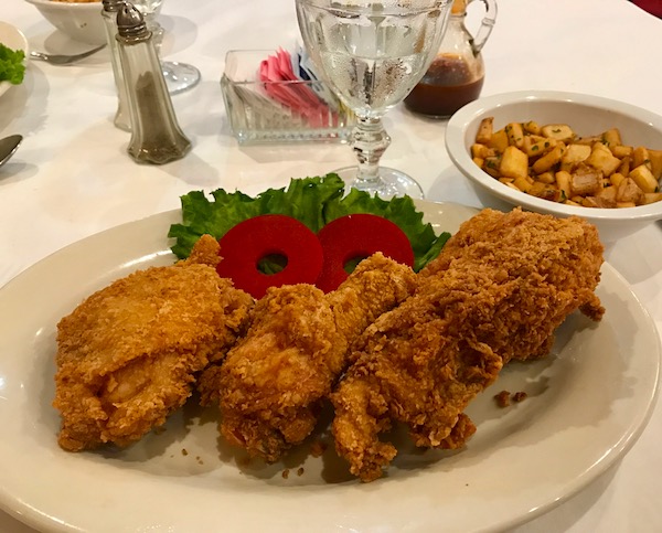 An ivory plate topped with fried chicken garnished with red apple rings and a lettuce leaf with a small bowl of roasted potatoes