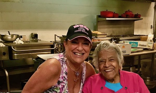 A woman in a ball cap leaning next to a woman in a hot pink chef coat with kitchen in background