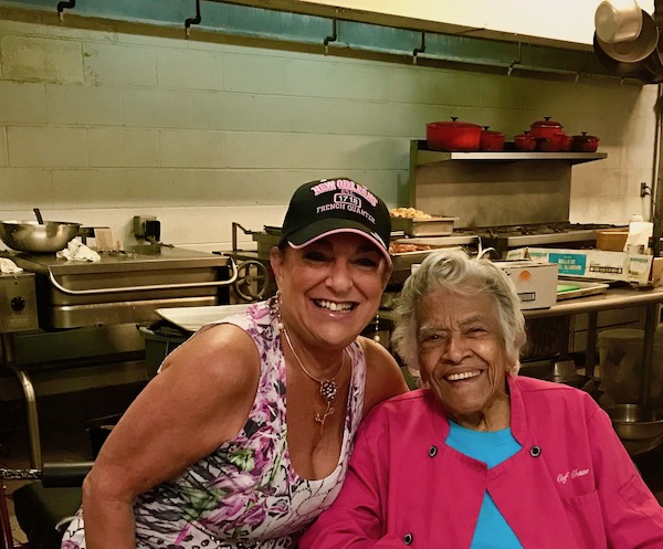 A woman in a ball cap leaning next to a woman in a hot pink chef coat with kitchen in background